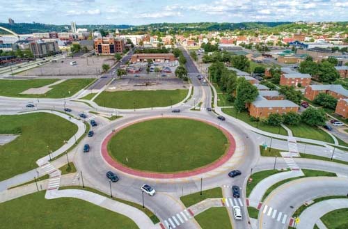 Aerial view of a traffic roundabout seamlessly integrated into urban road design, surrounded by cars, green spaces, and buildings in the background.