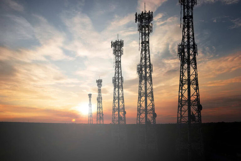 Five telecommunications towers stand in a row against a sunset sky with clouds, silhouetted above a dark landscape.