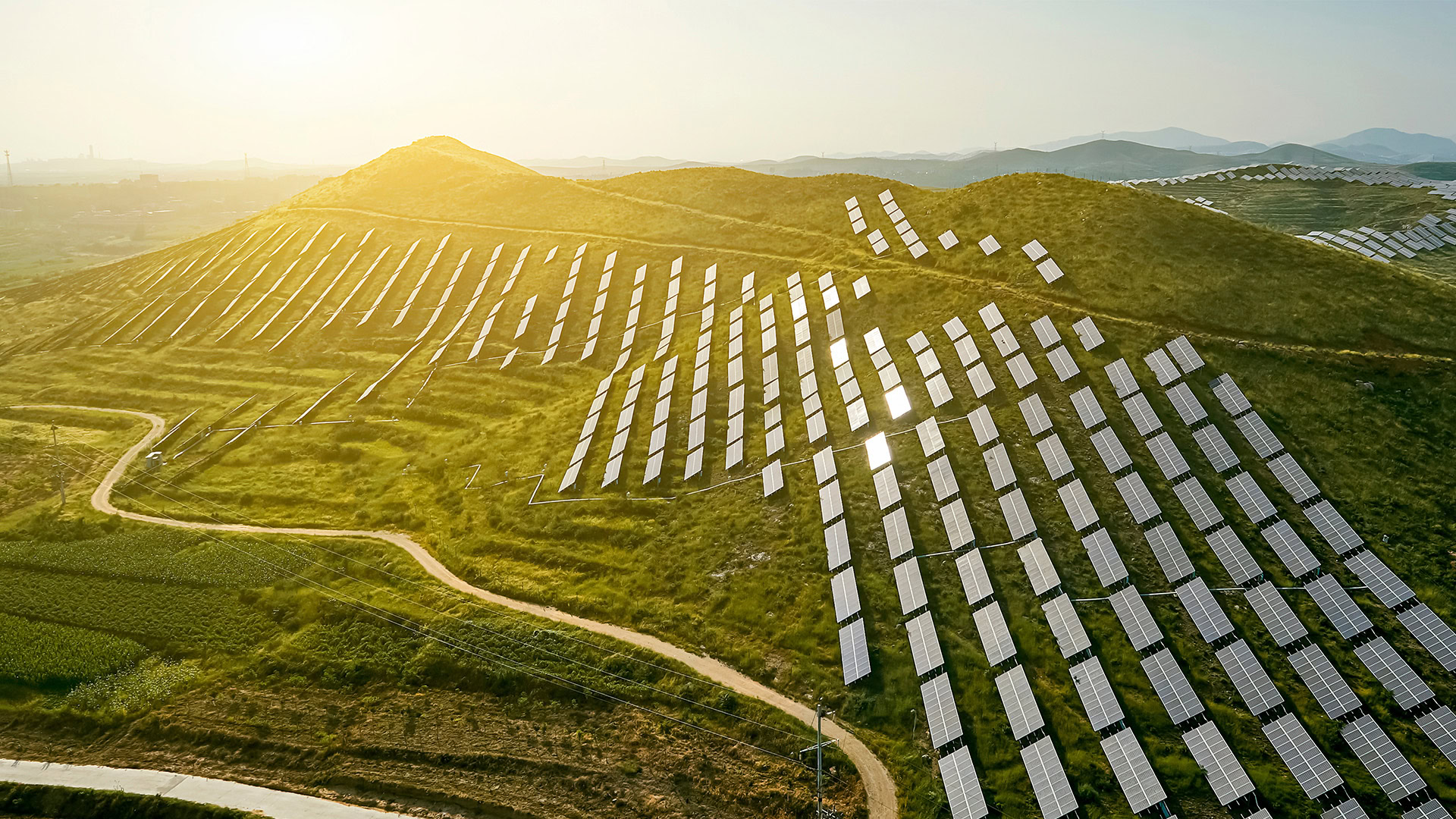 Aerial view of a solar farm on grassy hills, with rows of solar panels reflecting sunlight. A winding path cuts through the landscape.