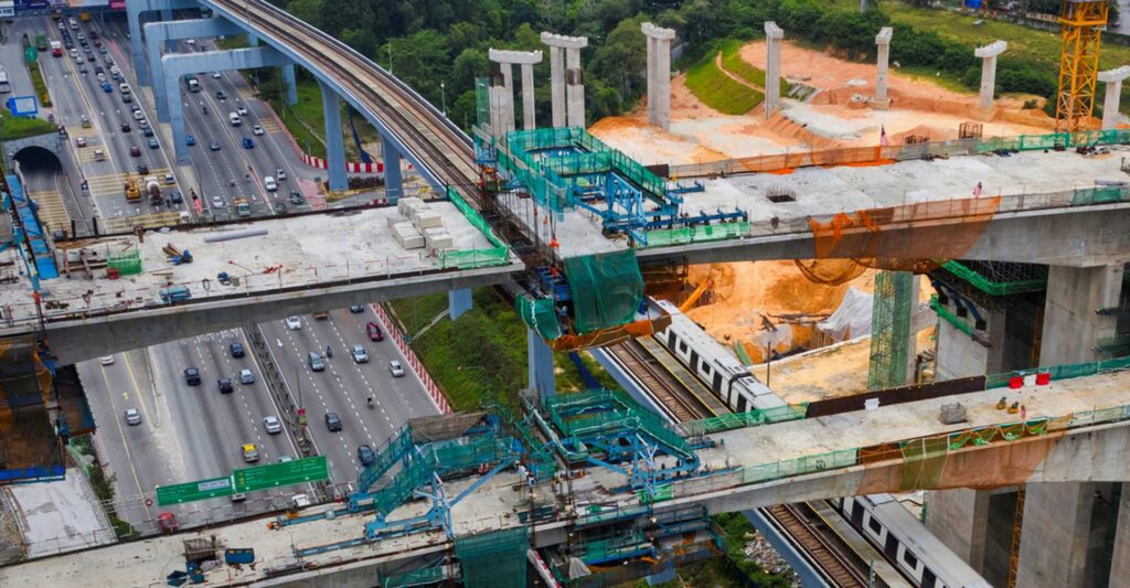 Aerial view of a highway next to a train track under construction with multiple elevated concrete structures and scaffolding. Vehicles are visible on the highway, surrounded by greenery. | Bentley Systems | Infrastructure Engineering Software Company Aerial view of a highway next to a train track under construction with multiple elevated concrete structures and scaffolding. Vehicles are visible on the highway, surrounded by greenery.