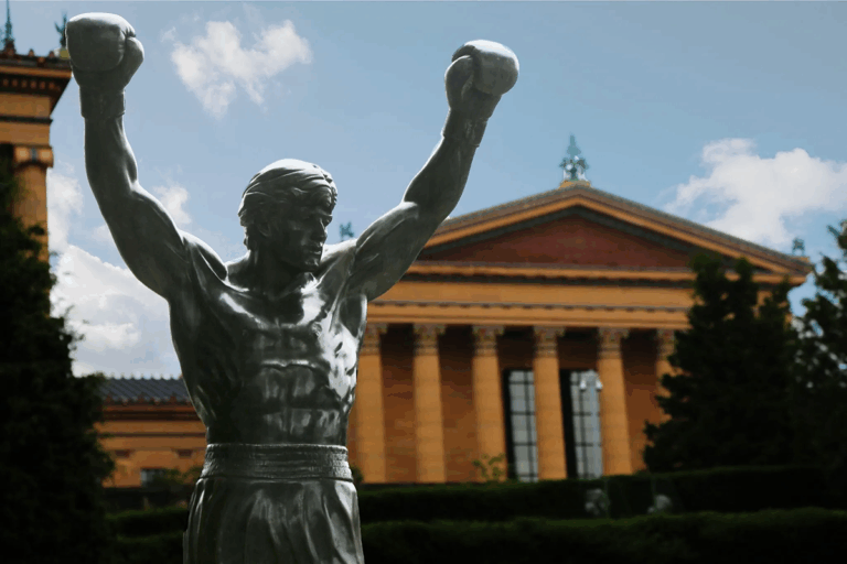 A bronze statue of a boxer with raised arms stands in front of a classical-style building with columns and a triangular pediment, making it a striking site for 3D geospatial demos at the dev conference.