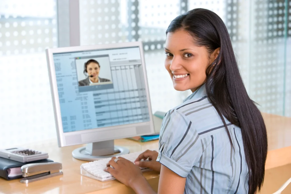 A woman sits at a desk, looking back with a smile while using Analysis Software on her computer, displaying a video call interface. | Bentley Systems | Infrastructure Engineering Software Company A woman sits at a desk, looking back with a smile while using Analysis Software on her computer, displaying a video call interface.