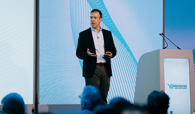 A person in a suit speaks on stage with a microphone. A podium is nearby and a large screen with abstract blue designs is in the background. Audience members are partially visible.